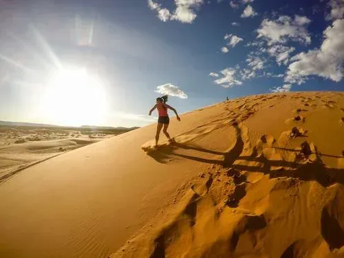 Person sandboarding down a sunlit desert dune under a blue sky