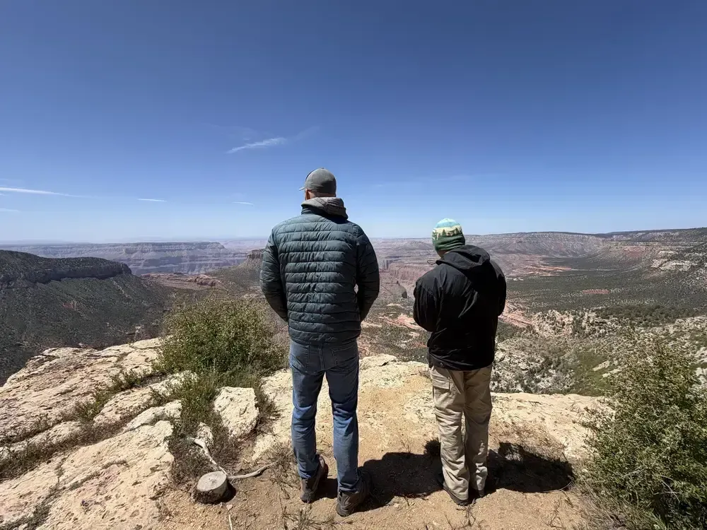Two people in jackets stand on a rocky overlook, facing a wide desert canyon under a clear blue sky.