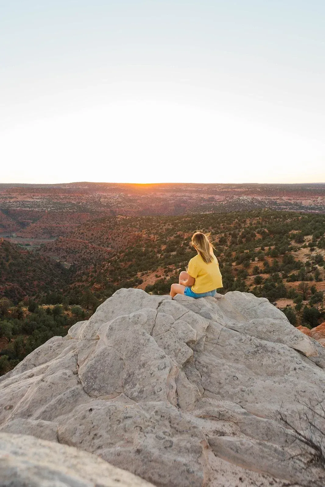 Person in a yellow shirt sitting on a rocky outcrop at sunset, overlooking a vast desert landscape.