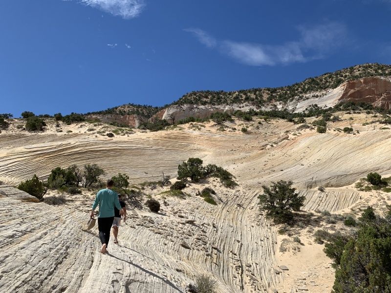 Two people hike on a striated sandstone surface towards a mountain under a blue sky.