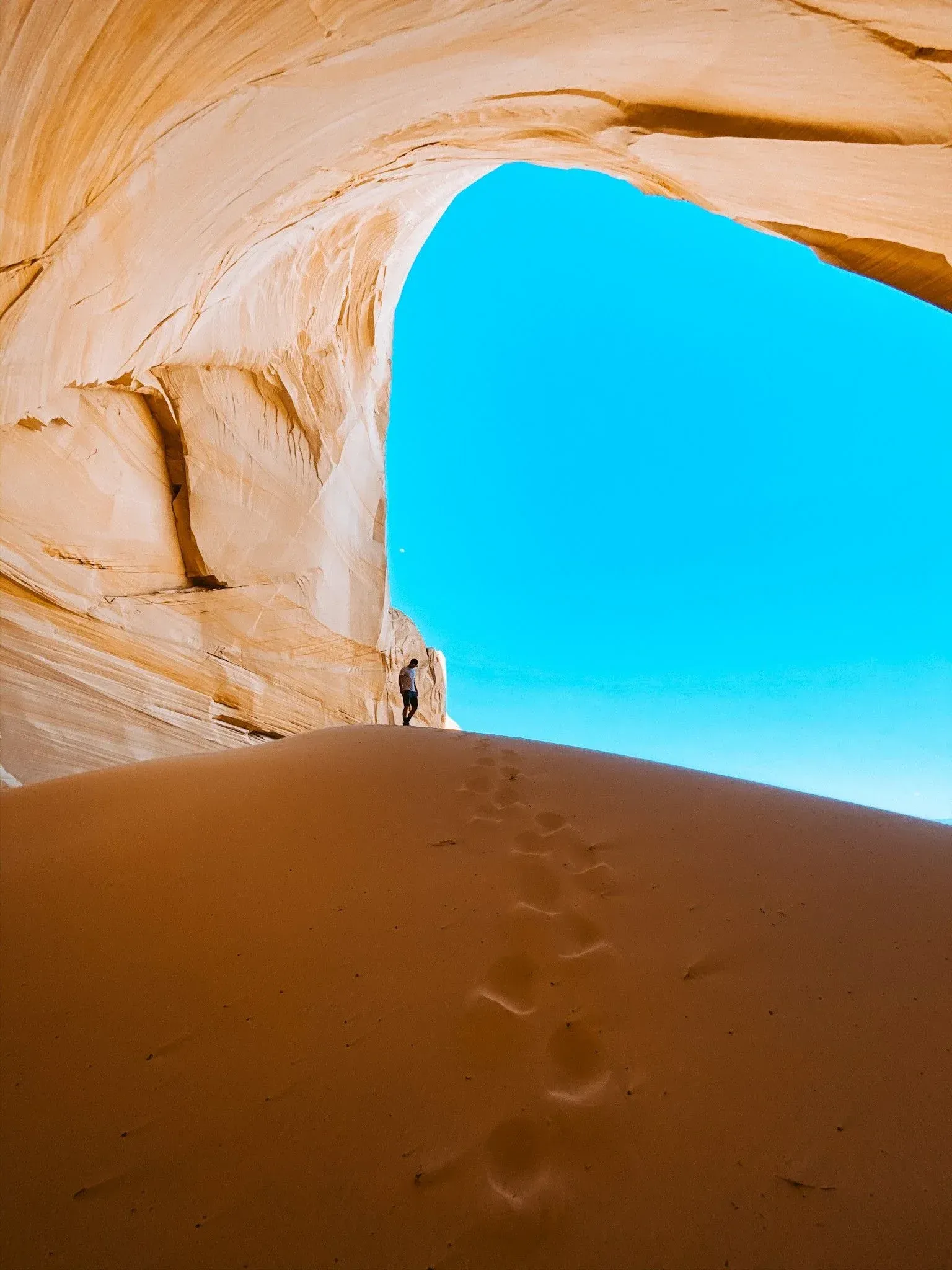 Footprints in a sand dune beneath a tan rock arch, with bright blue sky beyond