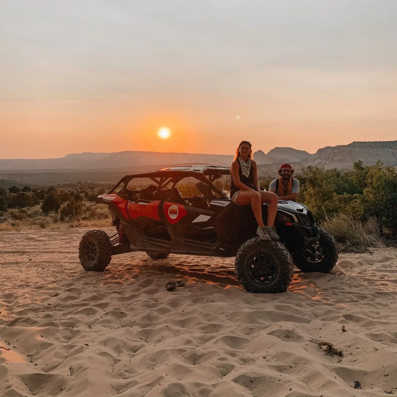 Two people on a red off-road buggy at sunset in a sandy desert landscape