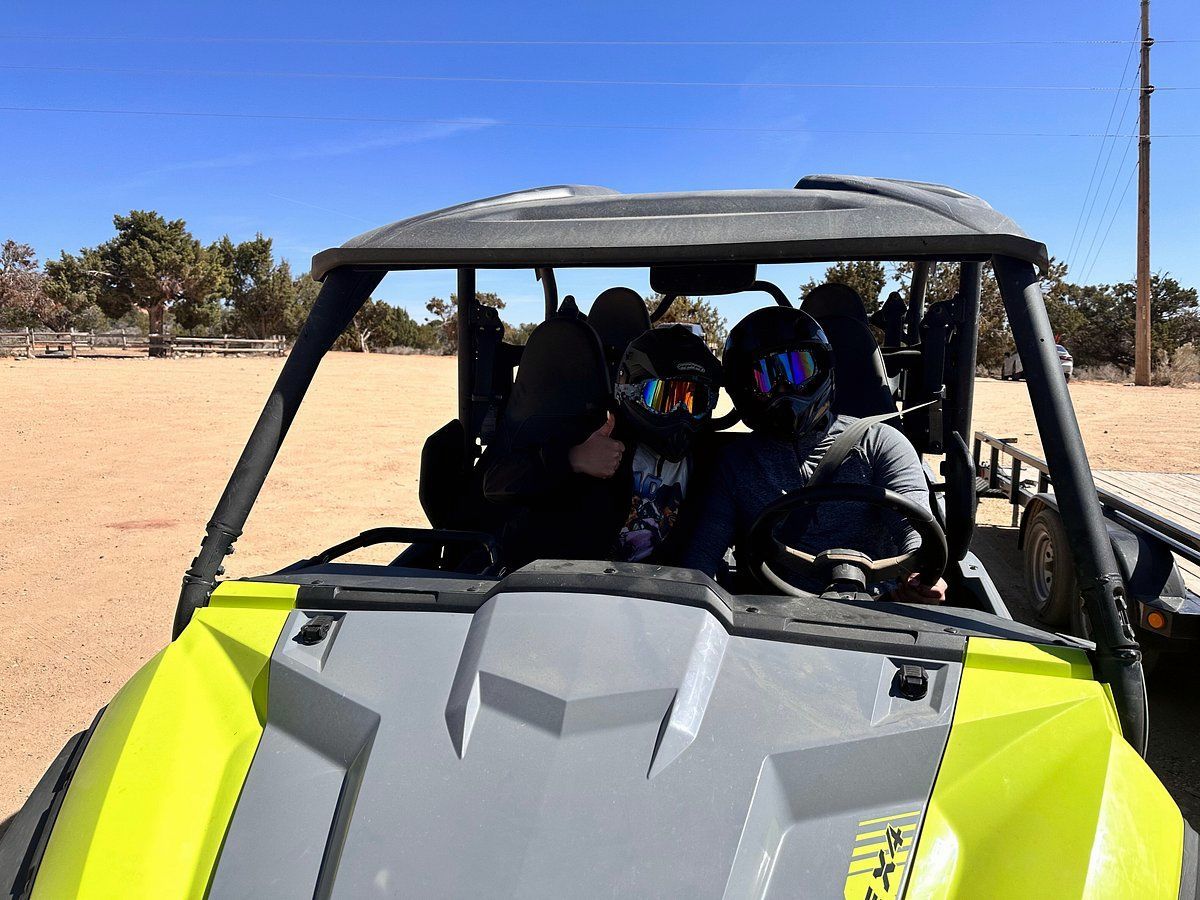 Two people in a gray off-road buggy driving on a sandy desert trail, wearing helmets.