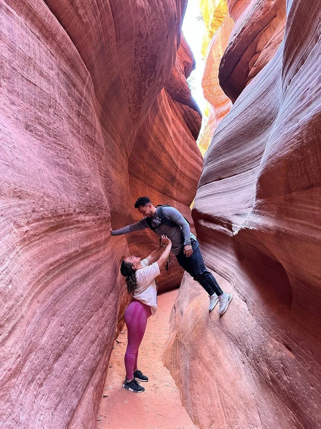 Two hikers climbing through a narrow red sandstone canyon, reaching for each other