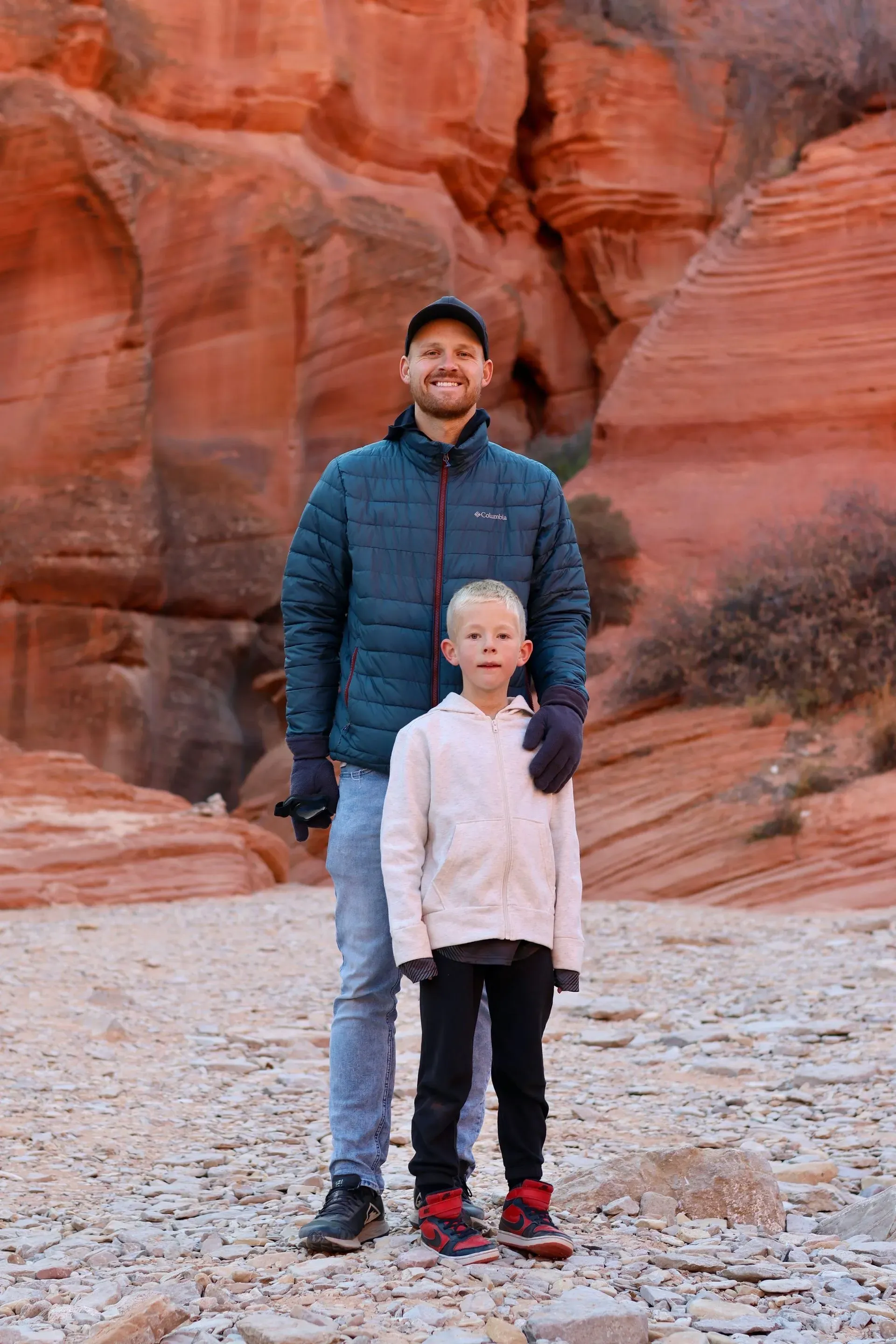 Two people standing on a rocky desert trail in front of red sandstone cliffs