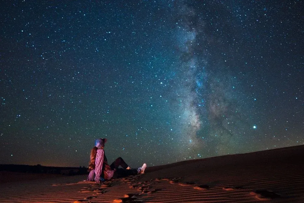 A person is sitting on top of a sand dune under a starry night sky.