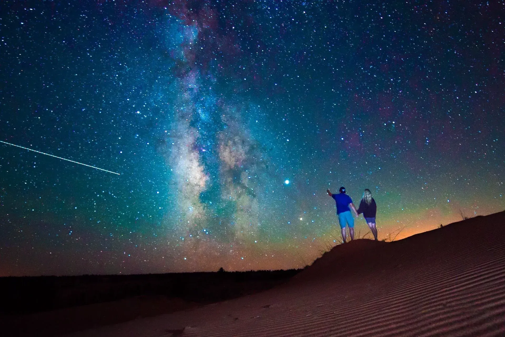 Two people on desert dunes under a vivid Milky Way night sky with a shooting star