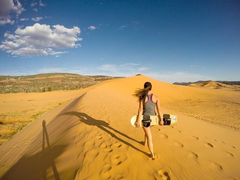 A woman is walking up a sand dune with a skateboard.
