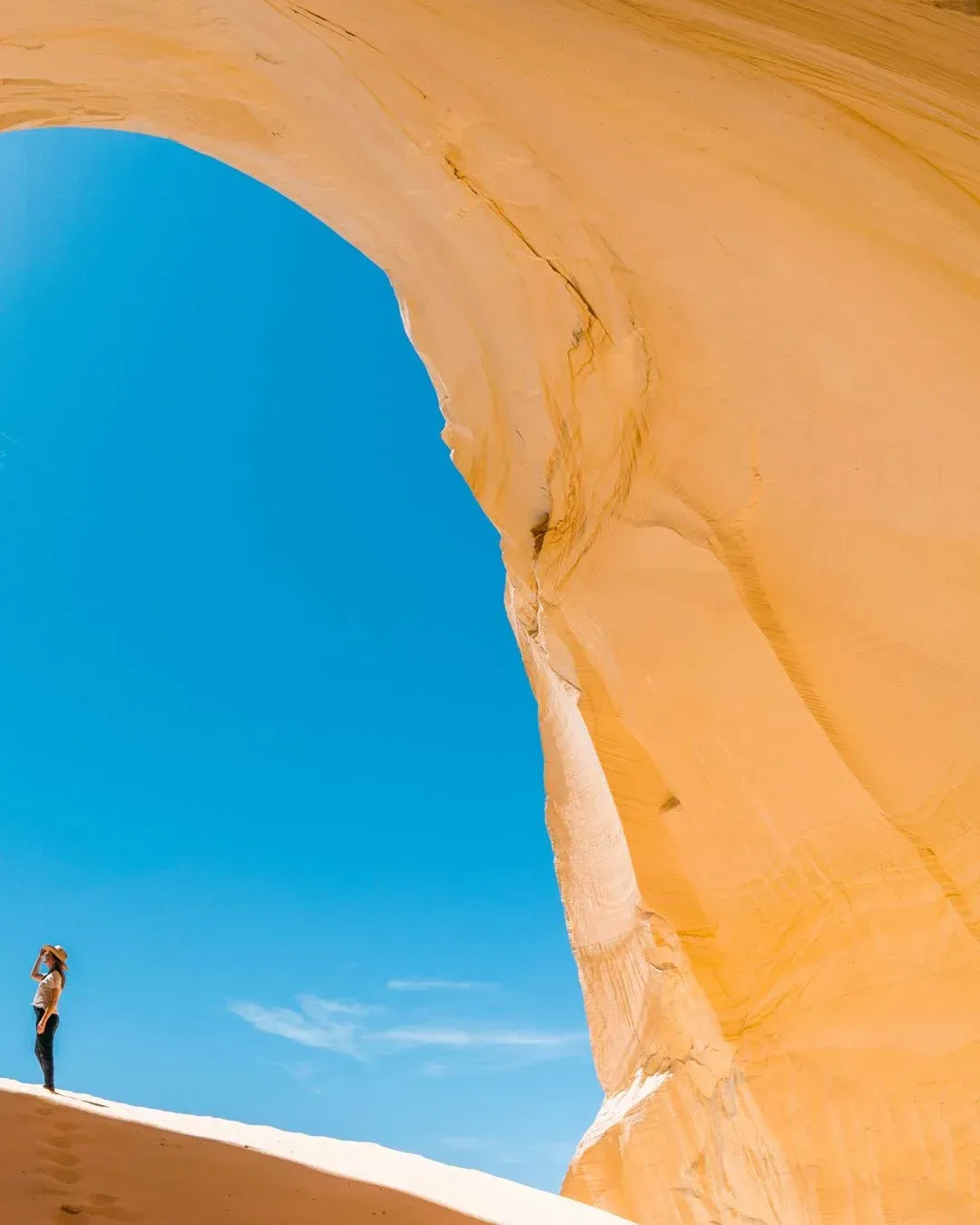 Person standing on white sand beneath a large orange rock arch against a bright blue sky