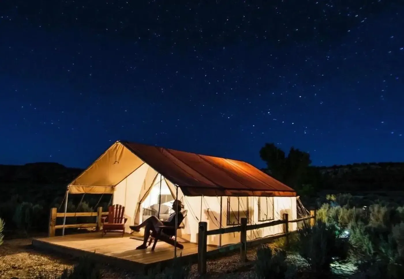Lit tent cabin on a wooden deck under a starry night sky in a rural field