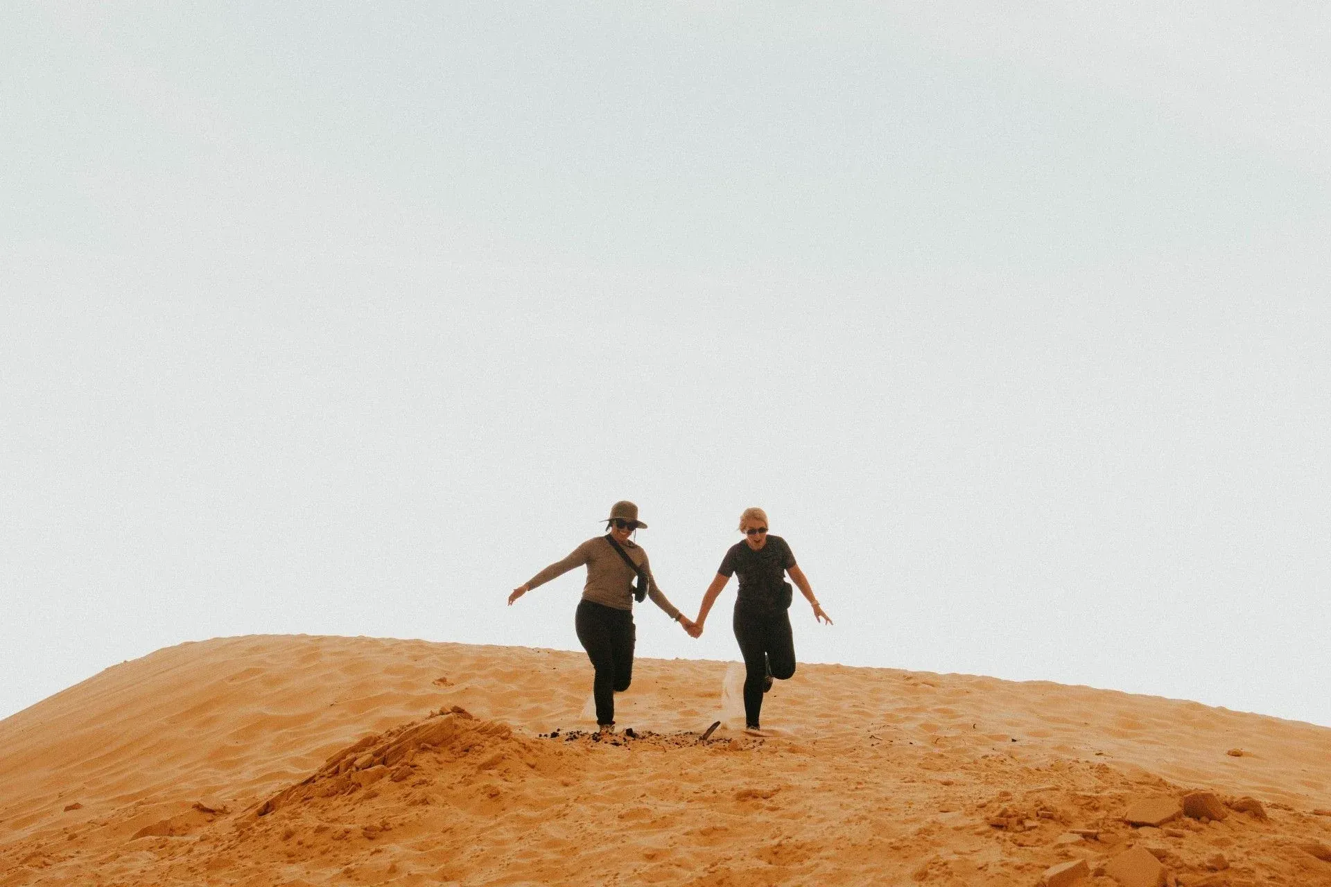 Two people holding hands, running over a brown sand dune against a pale sky.