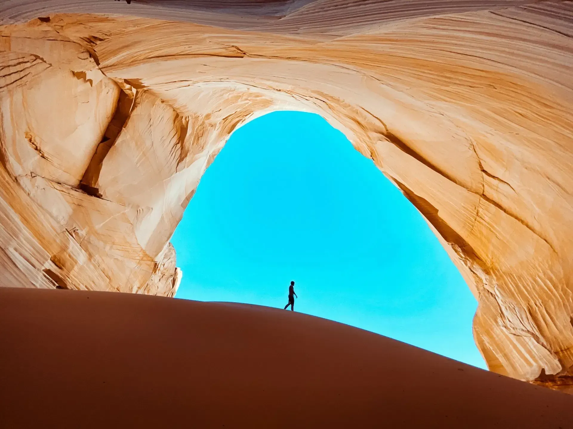 Silhouetted person under a sandstone arch framing bright blue sky and sand dunes