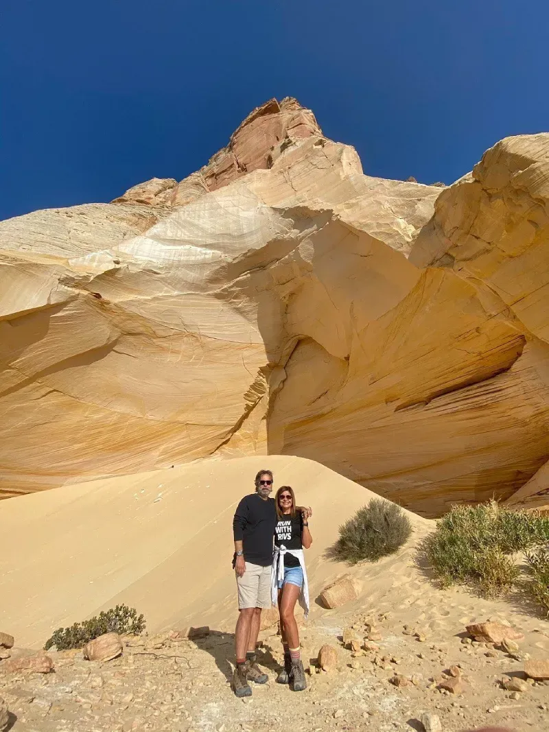 Two people standing on sandy ground beneath a sunlit tan rock formation under a blue sky