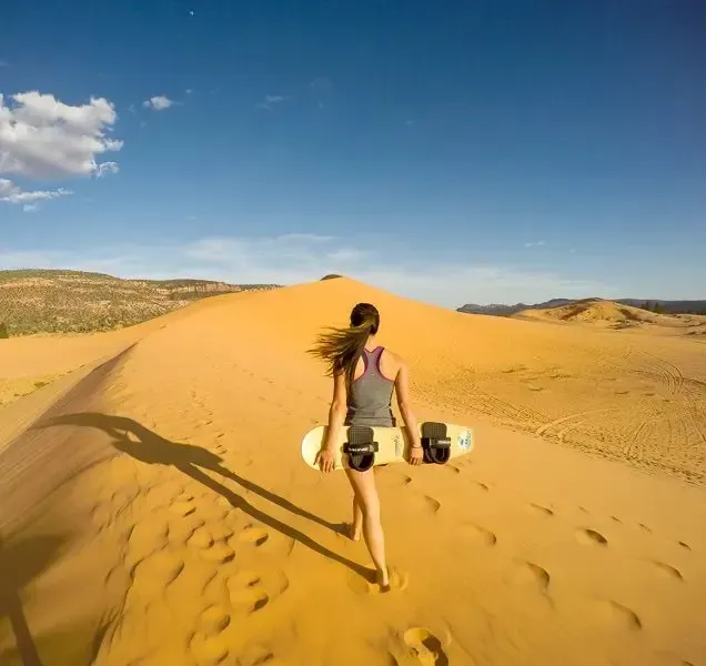 Person carrying a snowboard across golden sand dunes under a blue sky