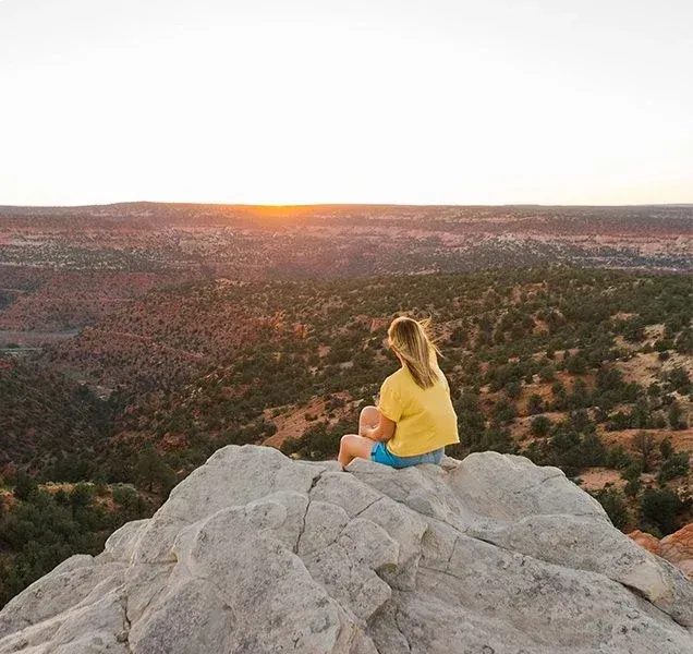 A woman is sitting on top of a rock looking at the sunset.