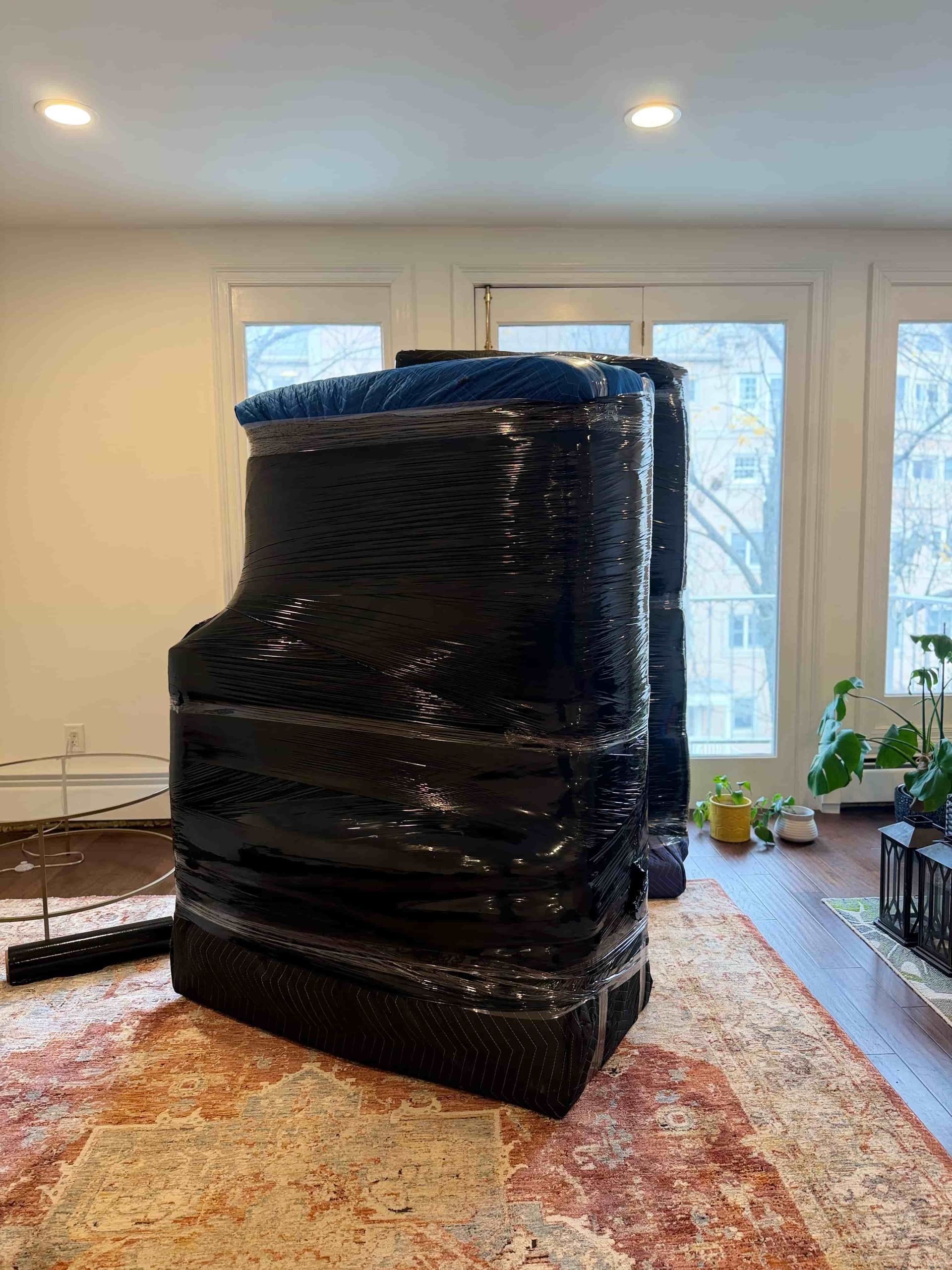 Wrapped piano, covered in black plastic, inside a room, near a window and rug.