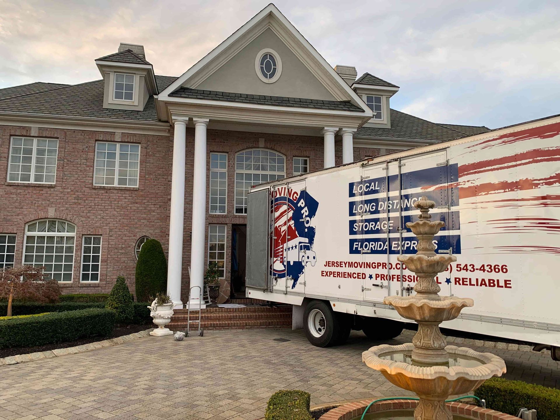 A large brick house with white pillars and a moving truck parked in the driveway, featuring a stone fountain in the front.