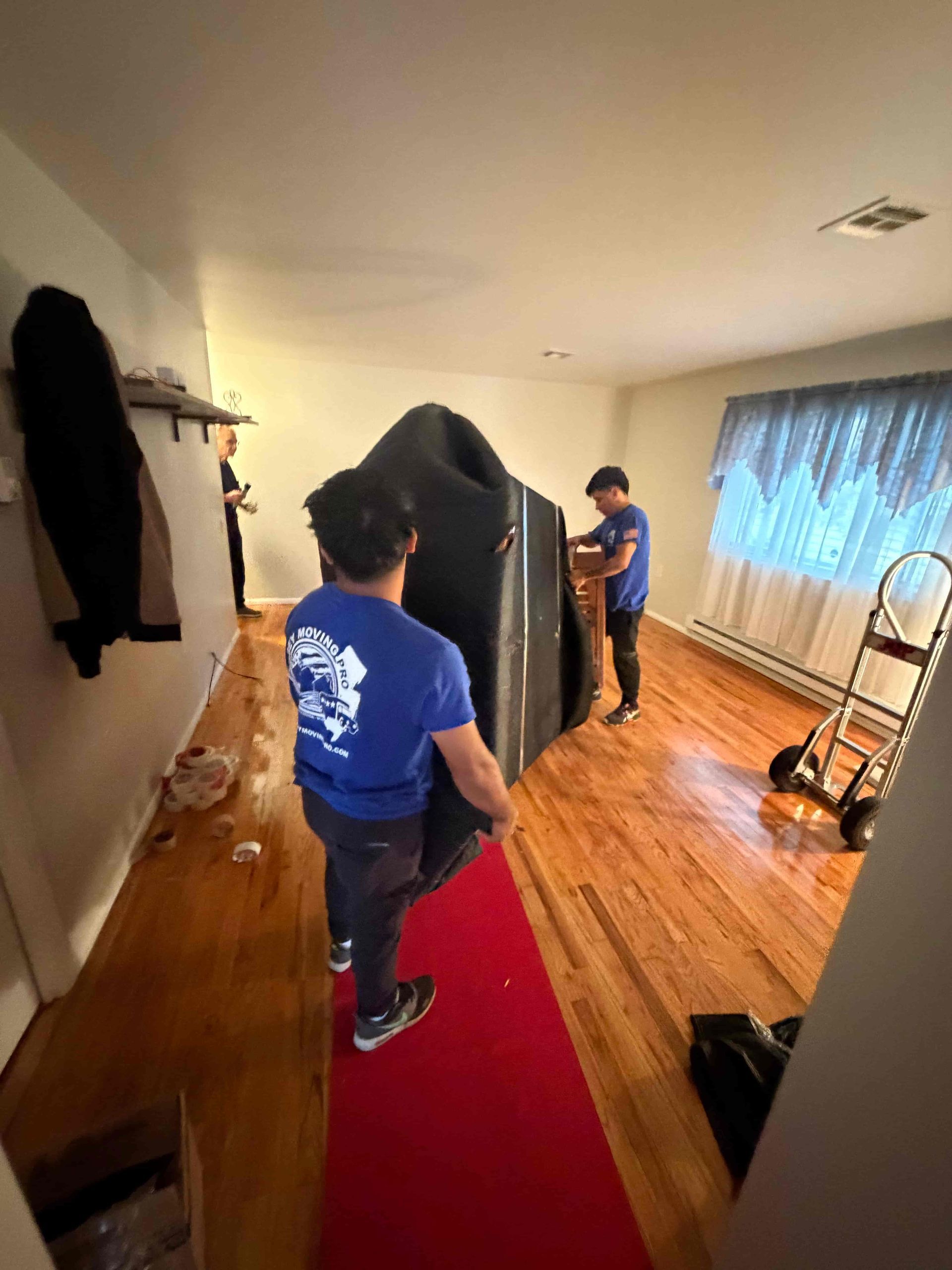 Three people moving a large, black object, possibly furniture, through a room with hardwood floors and red runner.