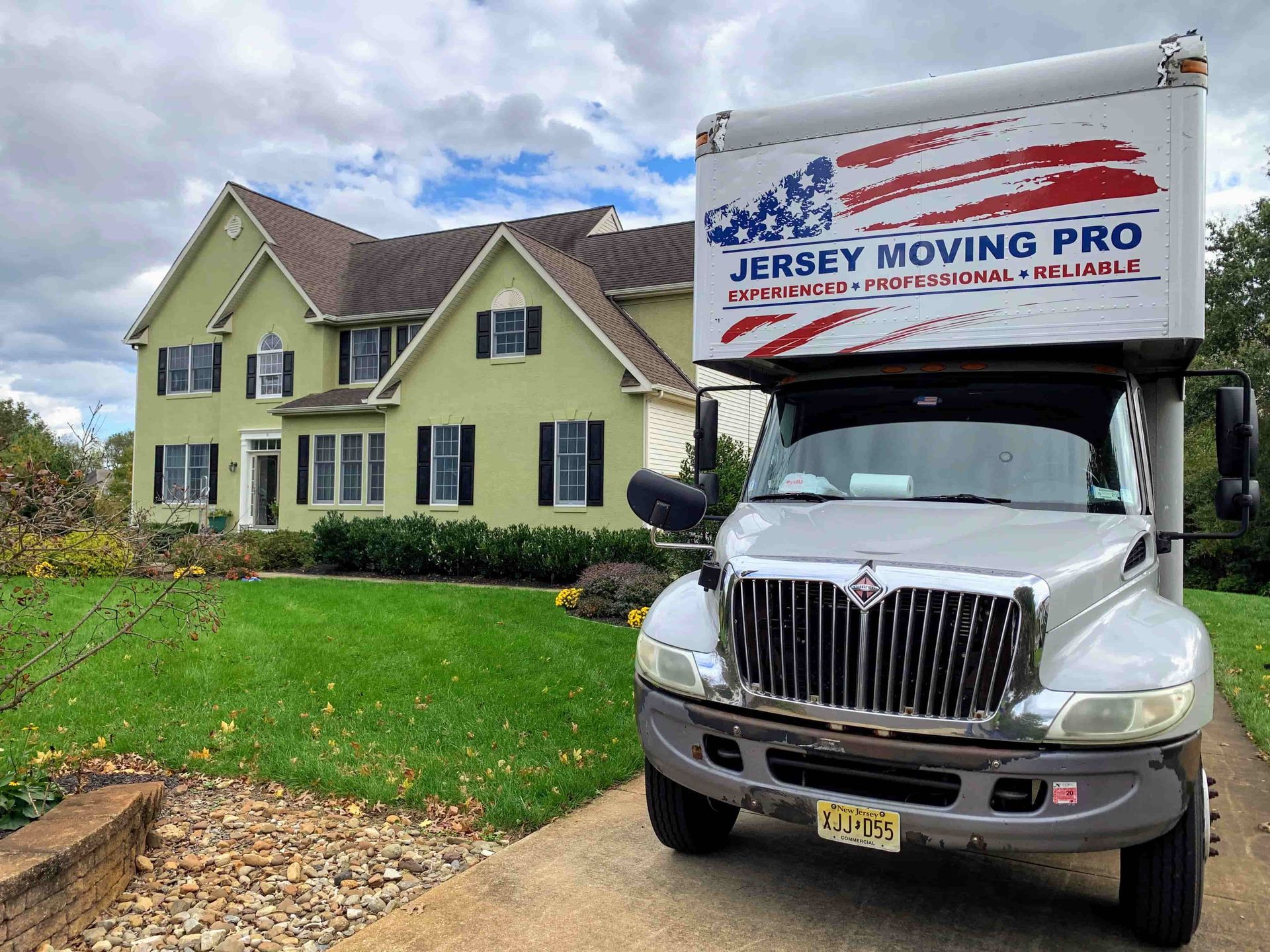 A Jersey Moving Pro truck parked in the driveway of a large, light green suburban house on a cloudy day.