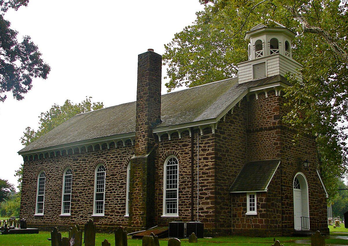 An old stone church with a white bell tower stands in a cemetery with green lawns and surrounding trees.