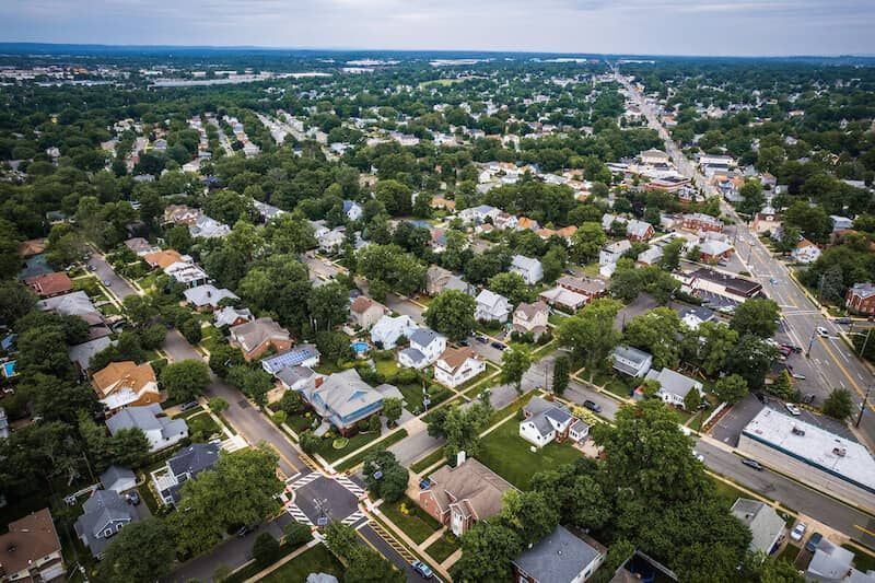 Aerial view of a suburban neighborhood with houses, streets, and trees; a road with shops and businesses is visible.