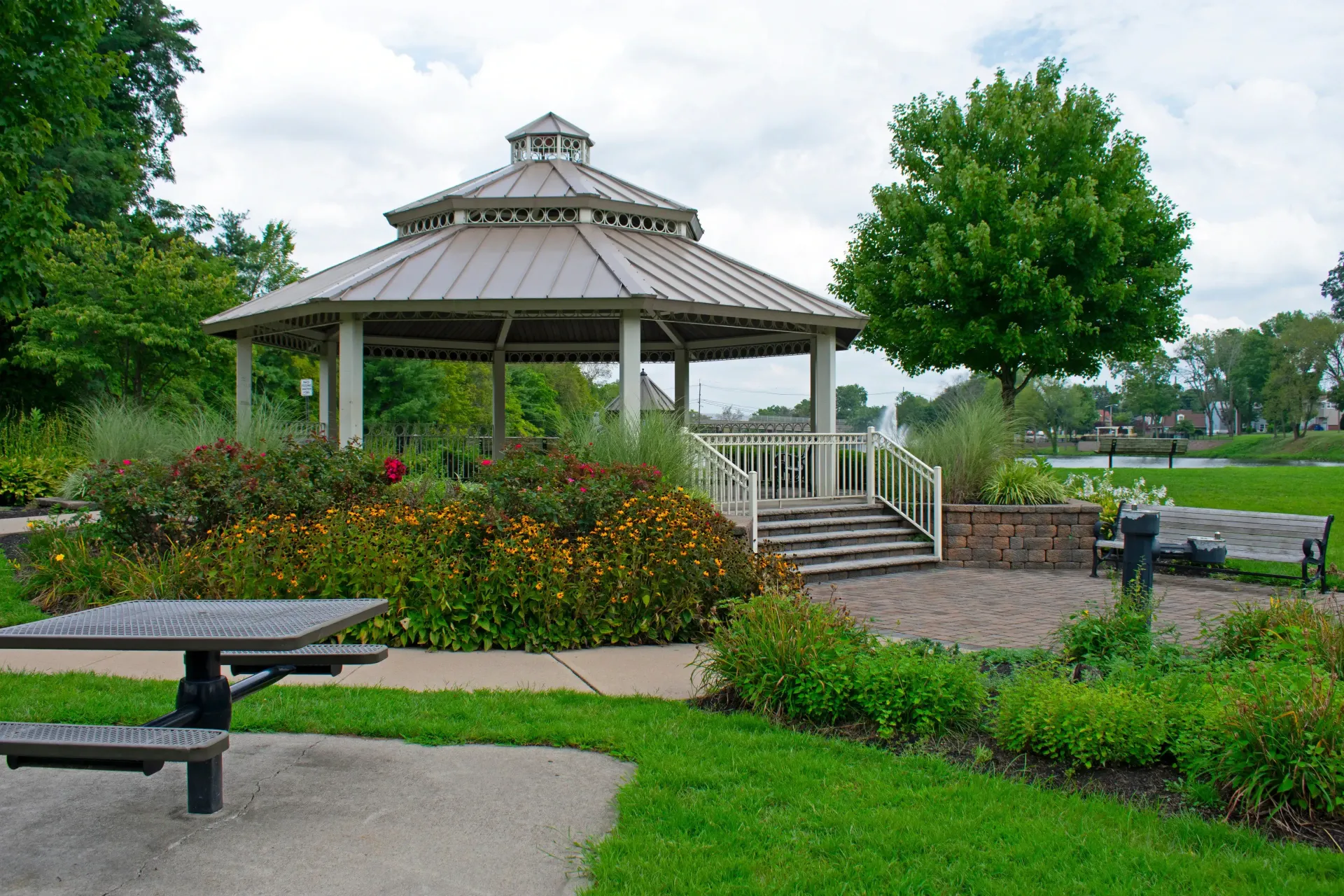 Gazebo in a park with flowers, picnic table, and benches. Green grass and trees surround. Overcast sky.