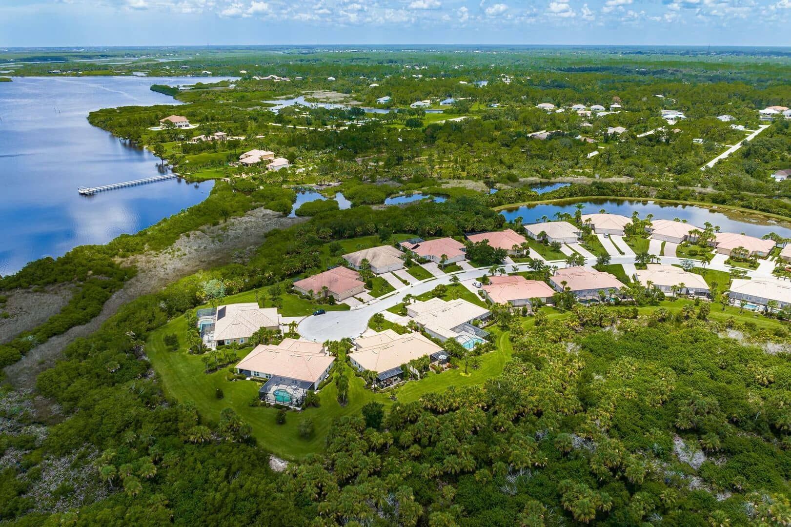 An aerial view of a residential complex with tile roofs nestled in a dense, green coastal landscape near blue water.