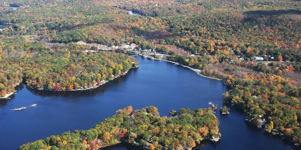 Aerial view of a deep blue lake surrounded by dense, colorful autumn forests with hints of small buildings on the shore.