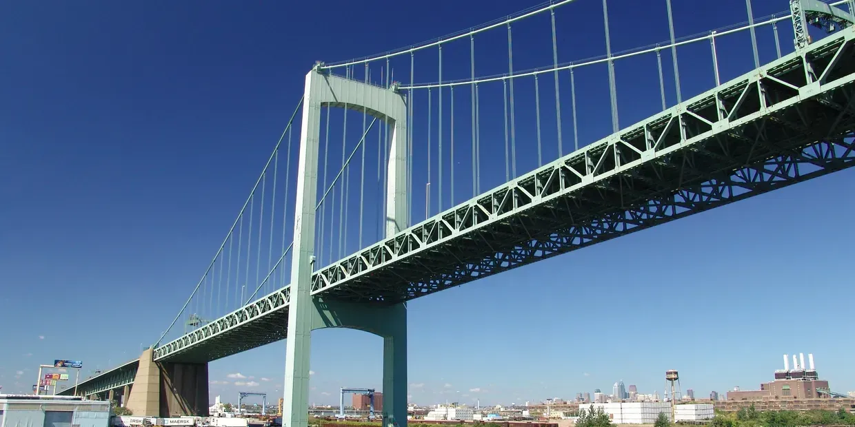 A green suspension bridge stretches across a clear blue sky above a city skyline.