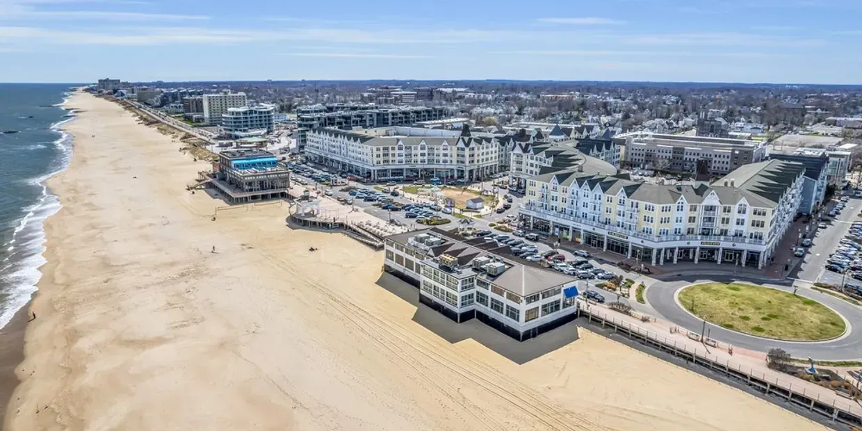 Aerial view of a beach with a pier, buildings, and a roundabout, under a blue sky.