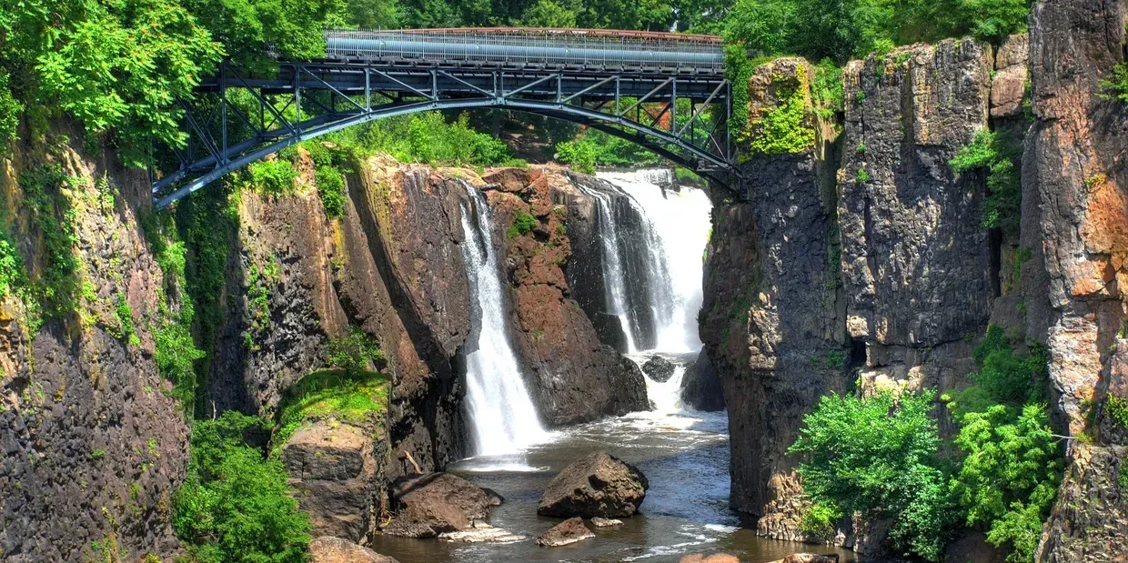 Waterfall cascading over rocks, a bridge arches above. Lush green trees frame the scene.
