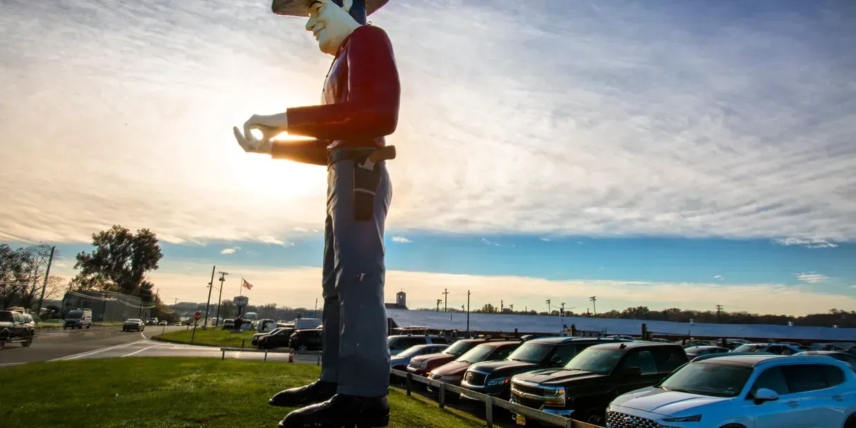 Giant cowboy statue, roadside. Red shirt, blue jeans, large parking lot with cars in background, sunny sky.