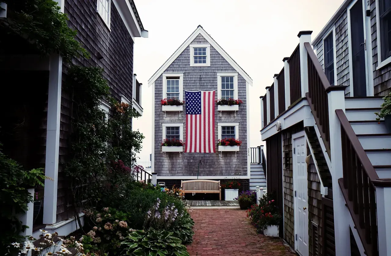 Narrow alley with brick path leads to a tall wooden house displaying a Betsy Ross flag, flanked by other buildings.