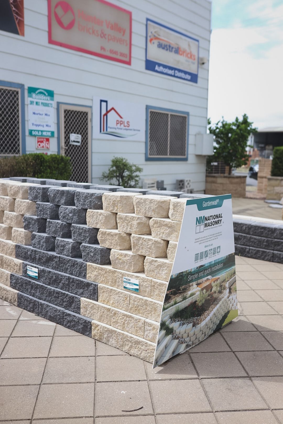 Stacked grey and beige stone blocks displayed outside a building on a tiled sidewalk.— Hunter Valley Bricks & Pavers in Scone, NSW