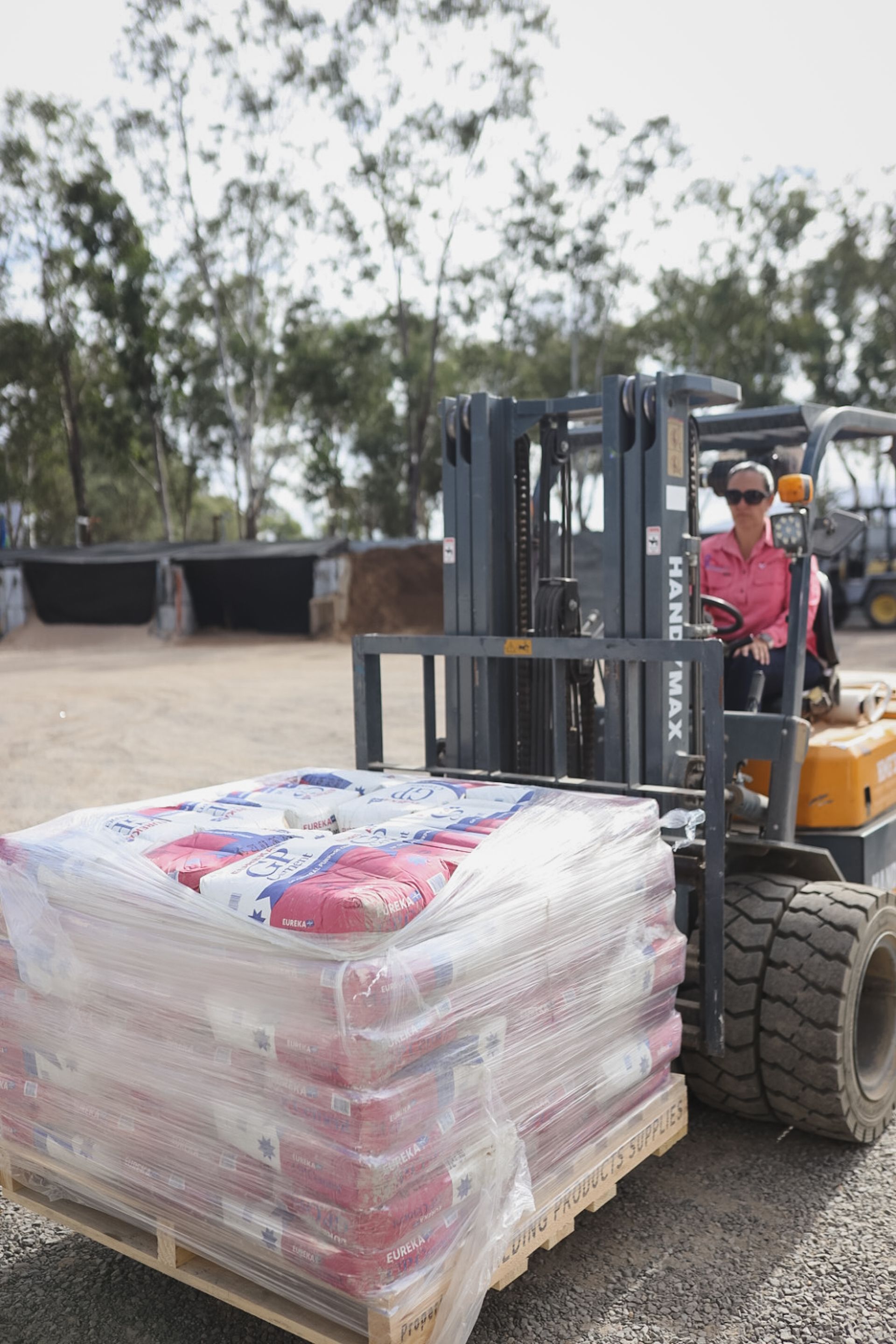 Forklift carrying a pallet of wrapped goods in an outdoor yard, with a worker nearby — Hunter Valley Bricks & Pavers in Scone, NSW
