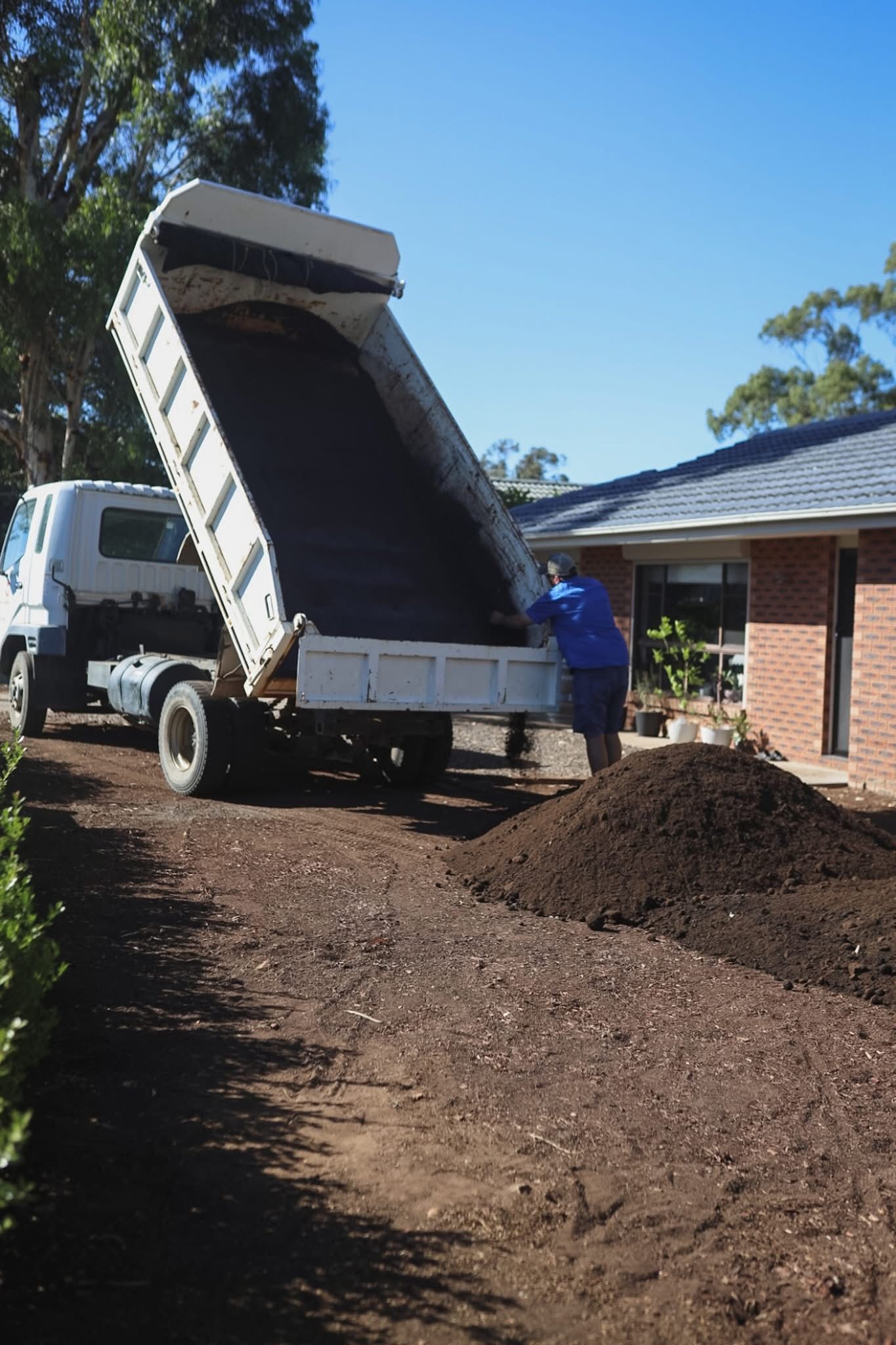 Dump truck unloading mulch beside a brick house, with a worker standing nearby.— Hunter Valley Bricks & Pavers in Scone, NSW
