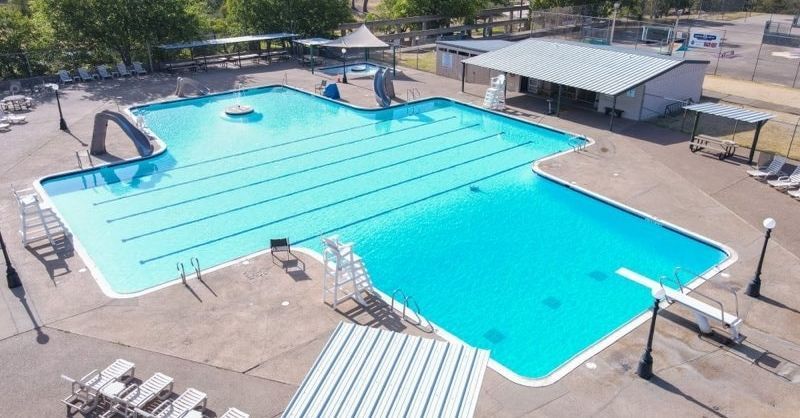 Aerial view of an empty outdoor swimming pool with blue water, diving board, and shaded deck area