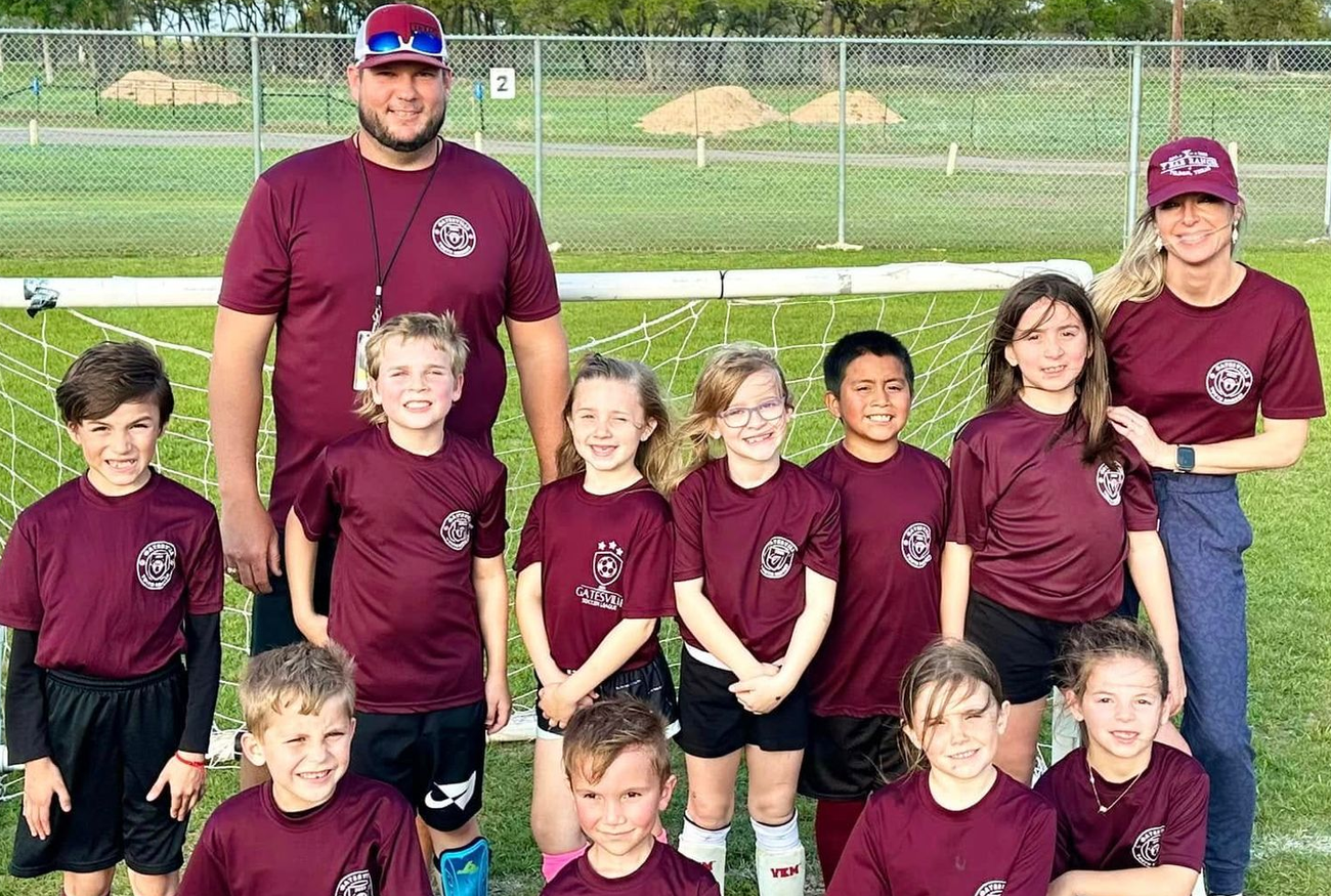 Youth baseball team posing with coach on a field in maroon jerseys; one girl holds a trophy.