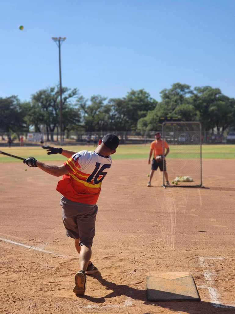 Softball batter in orange jersey swings at home plate while pitcher waits on dusty field.