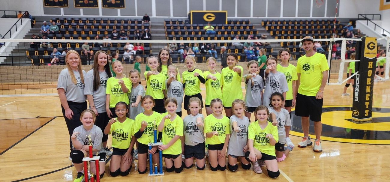 Youth volleyball team posing on an indoor court, wearing neon shirts with a volleyball net and bleachers behind them