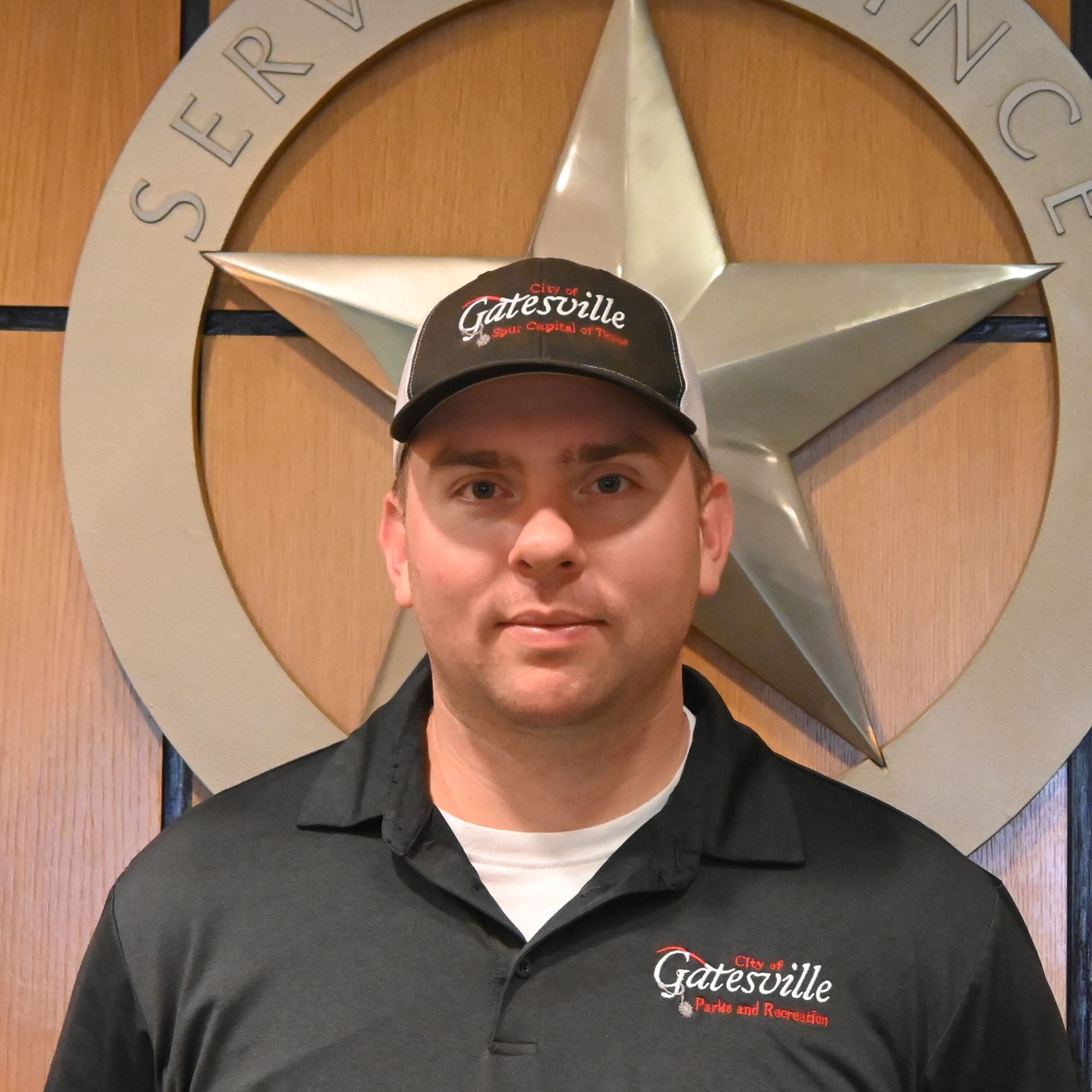 Man in black polo and cap standing before a large metal star wall emblem