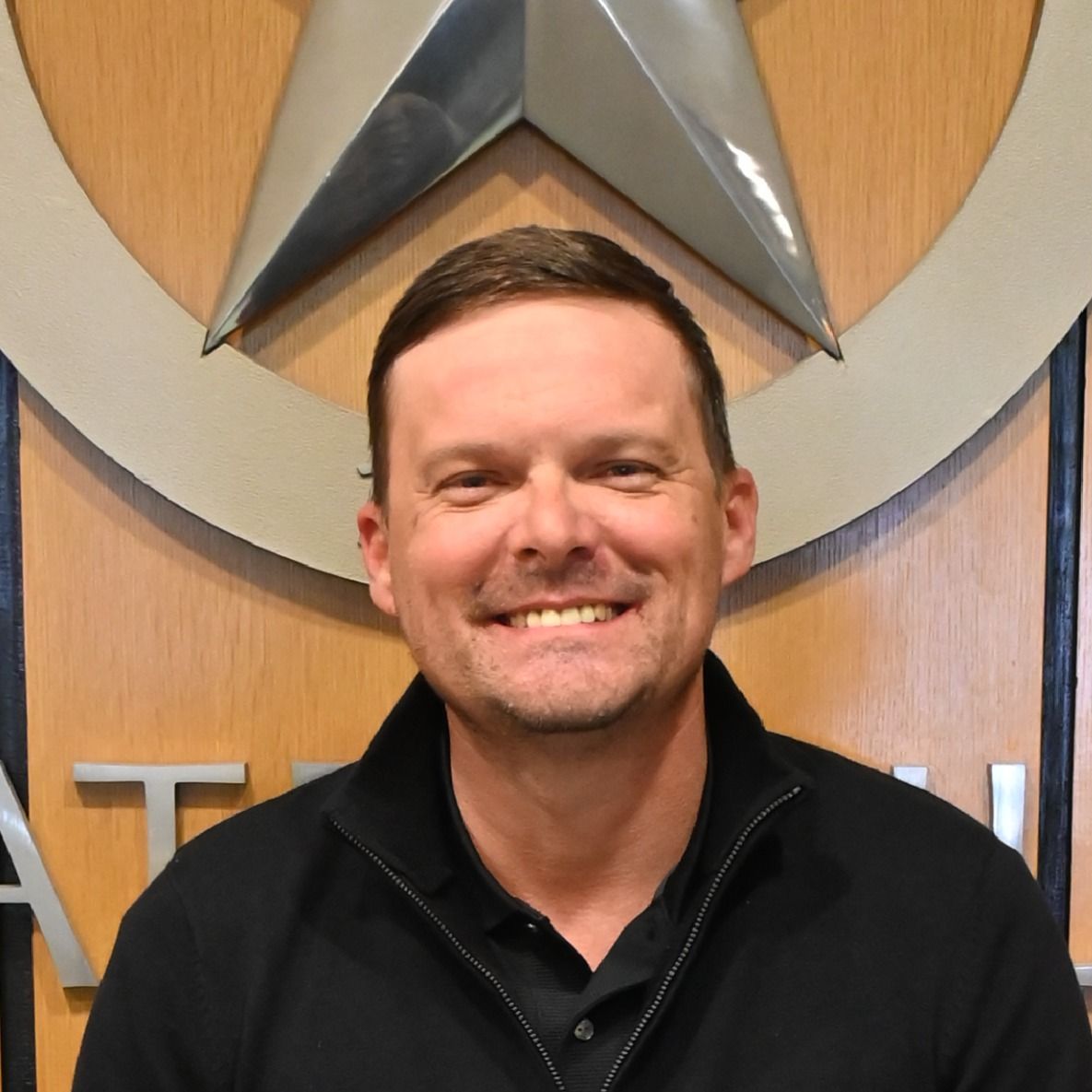 Smiling man in a black jacket standing in front of a wooden wall with a large star emblem