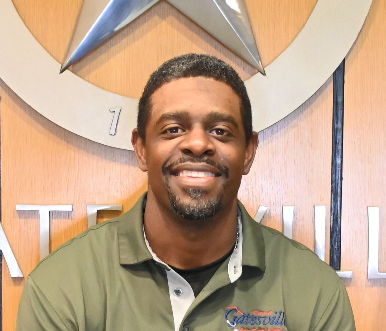 Smiling man in a green collared shirt standing before a wood-paneled wall with a silver star logo