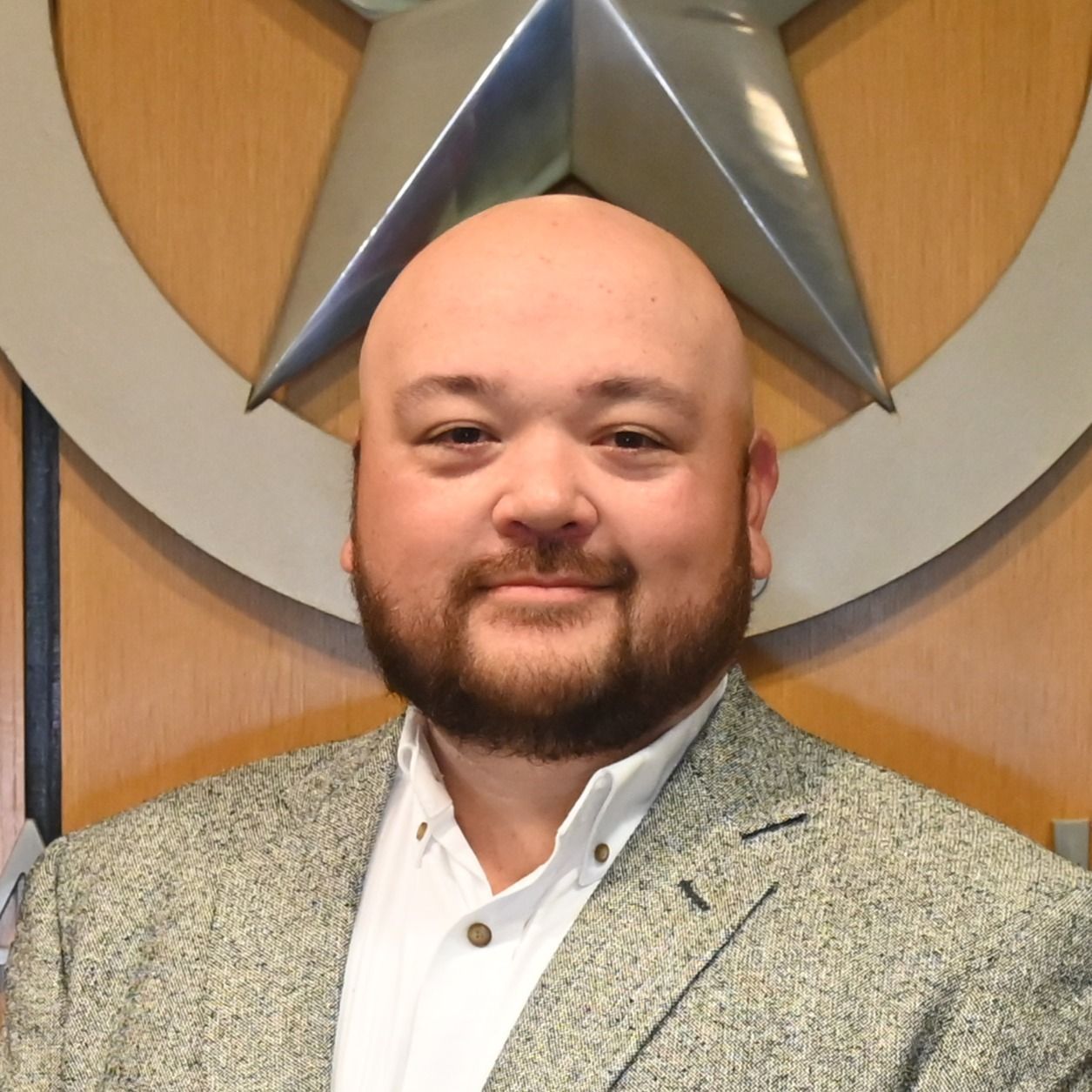 Smiling bald man in a gray blazer posing before a large circular emblem