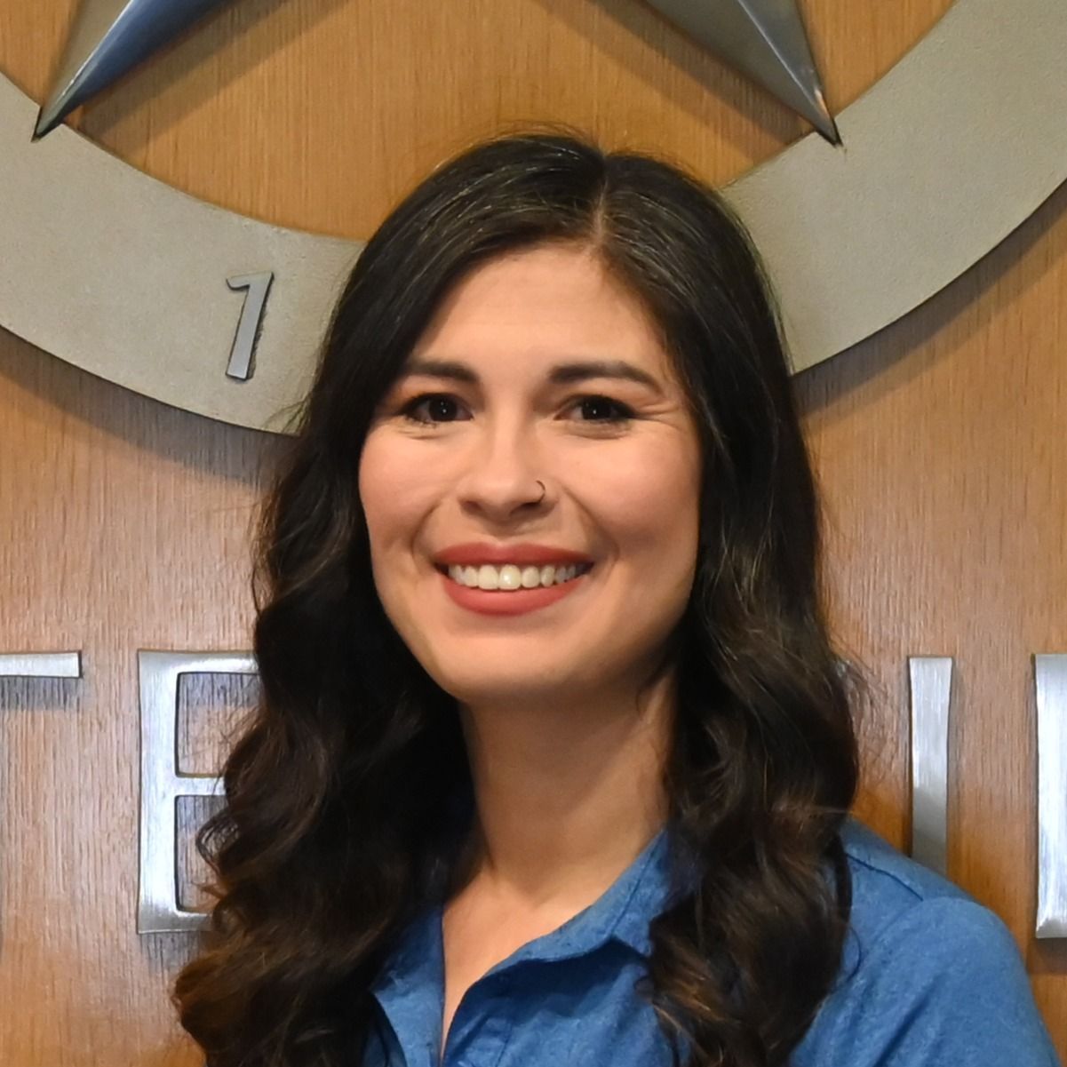 Smiling woman in a blue shirt standing before a wooden wall with a large clock.