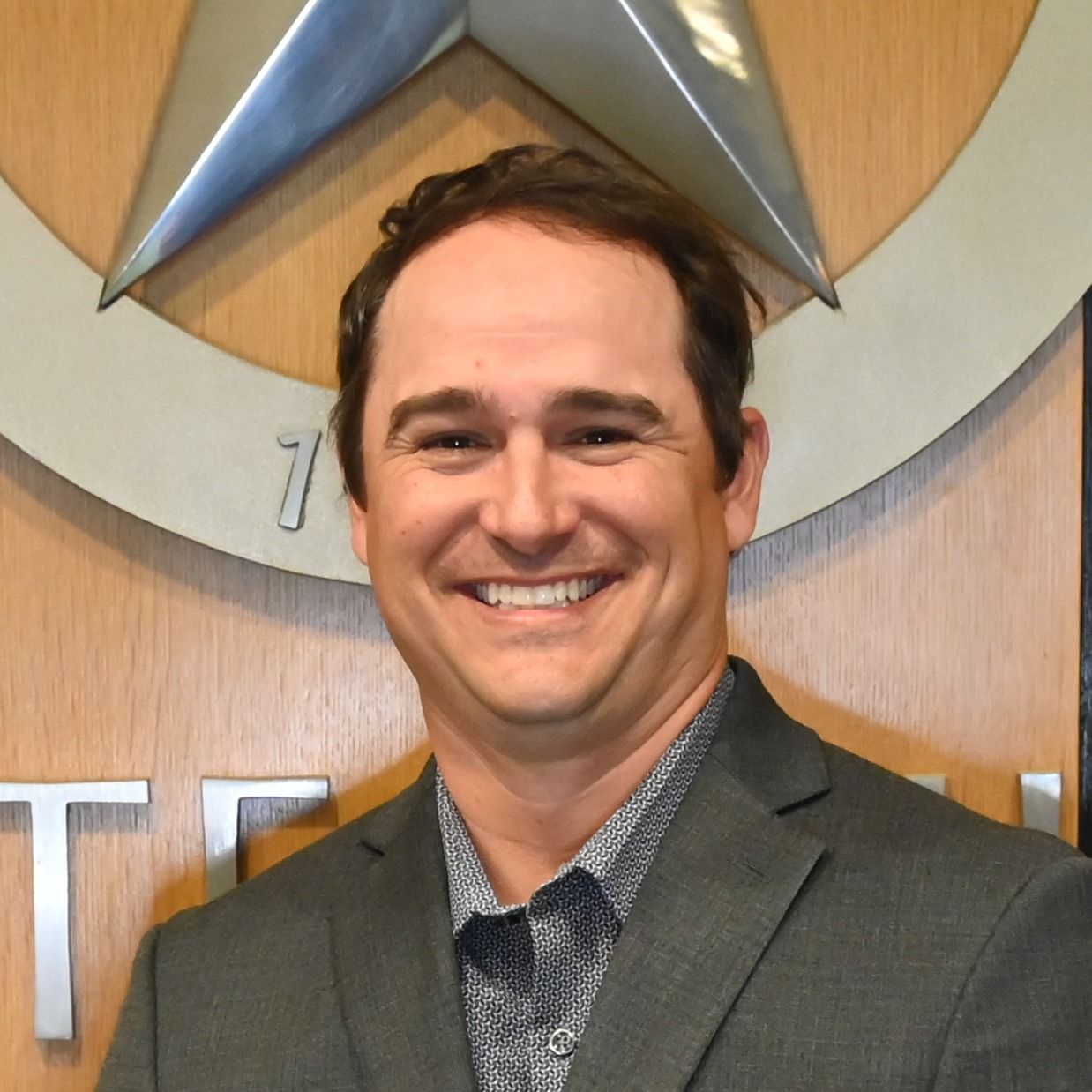 Smiling man in a gray blazer standing before a large star emblem on a wood wall