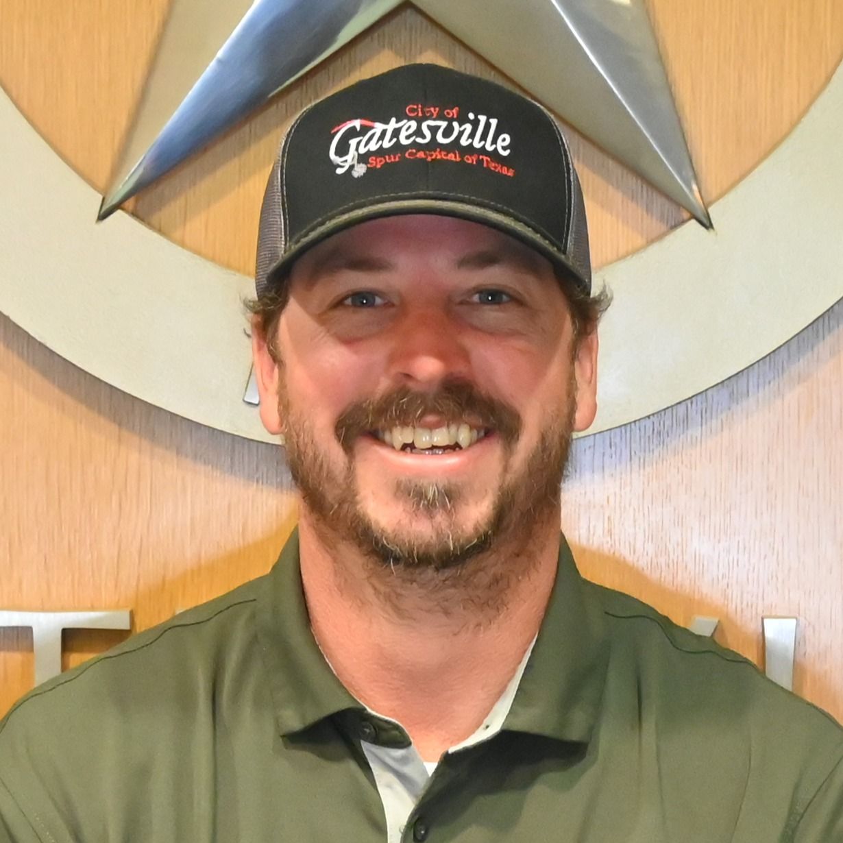 Smiling man in a green shirt and black cap standing before a logo wall