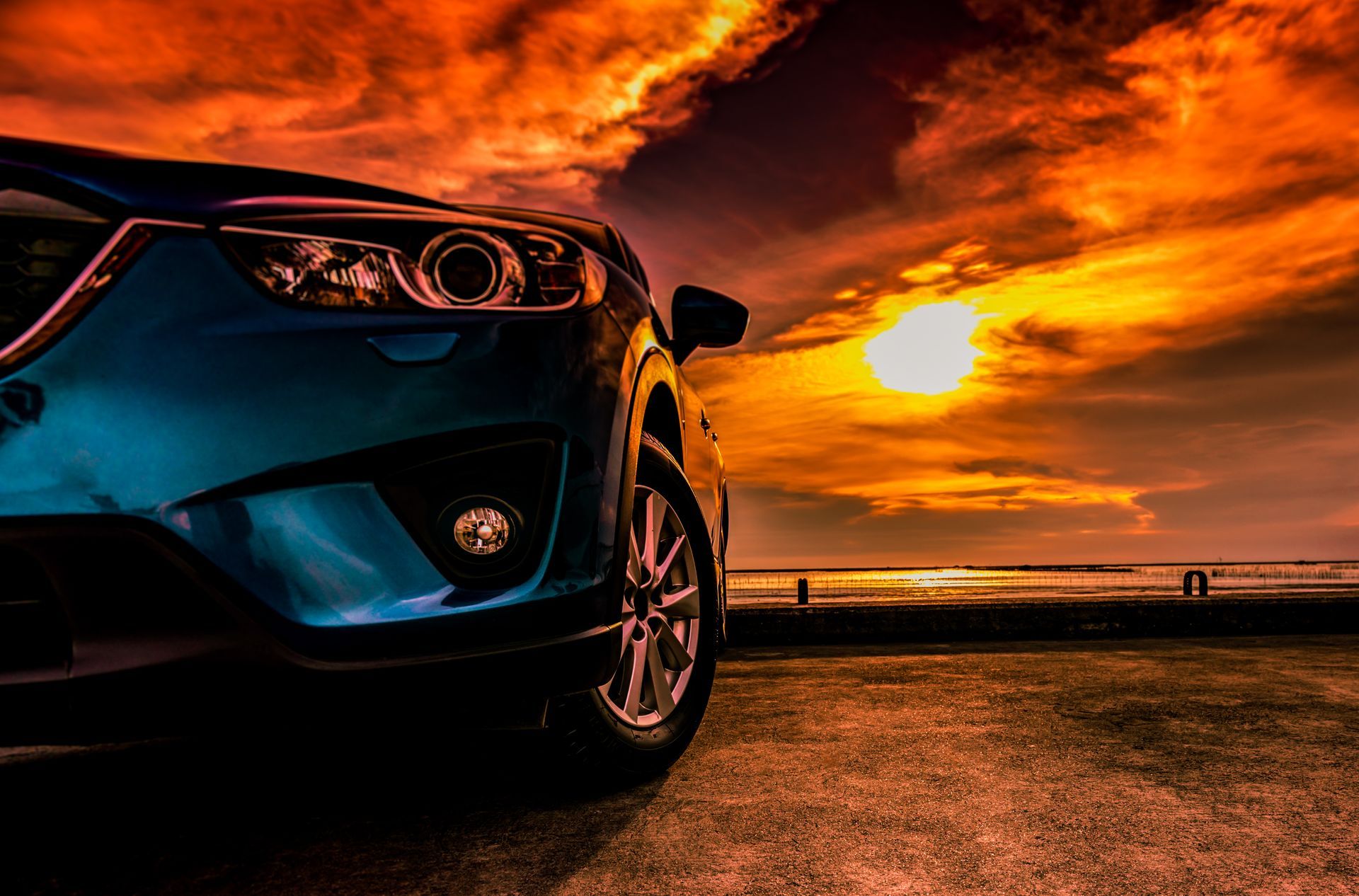 Blue SUV parked on a beach at sunset; fiery orange sky reflects on the car's hood.