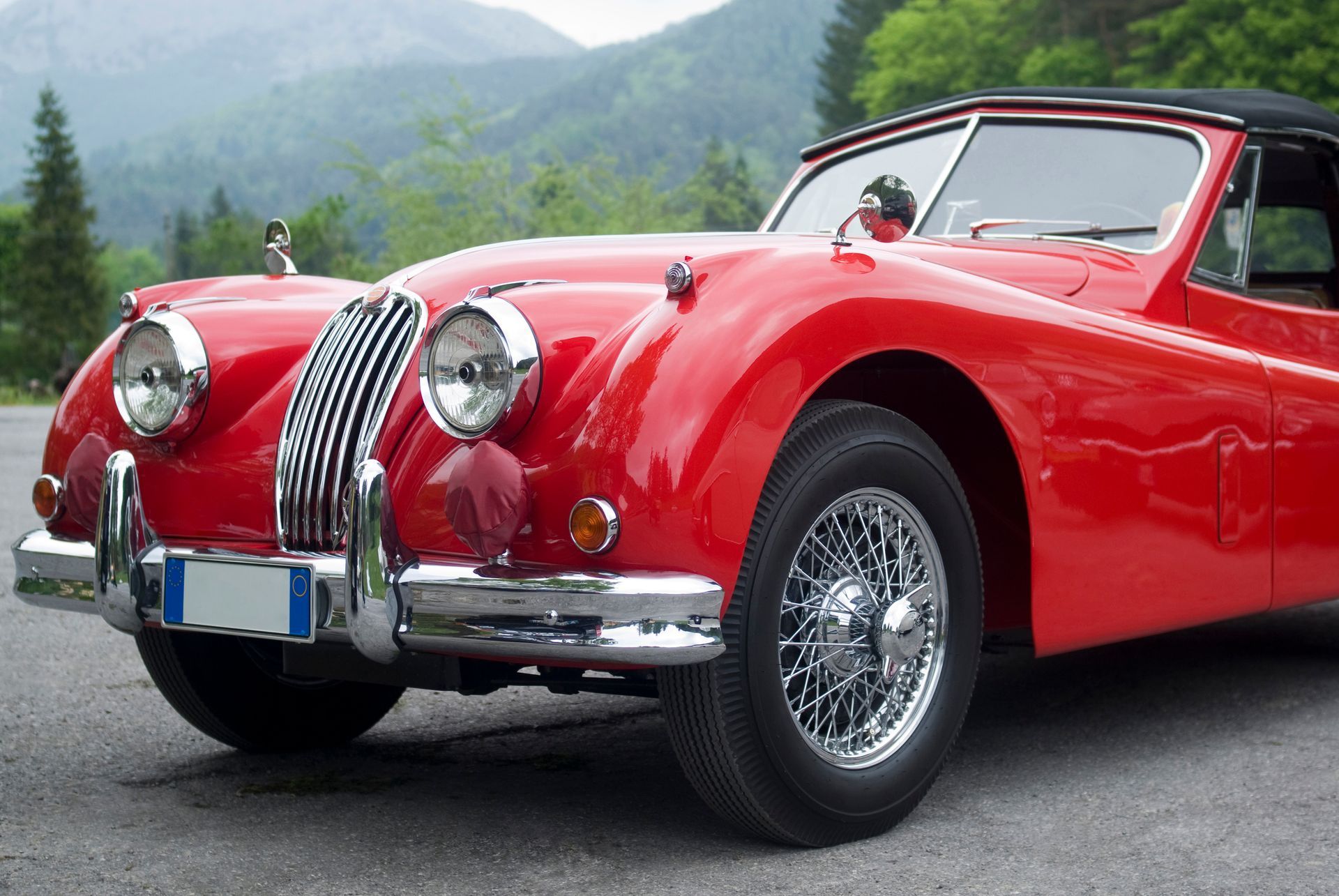 Red vintage Jaguar convertible with wire wheels, parked in front of a mountain backdrop.