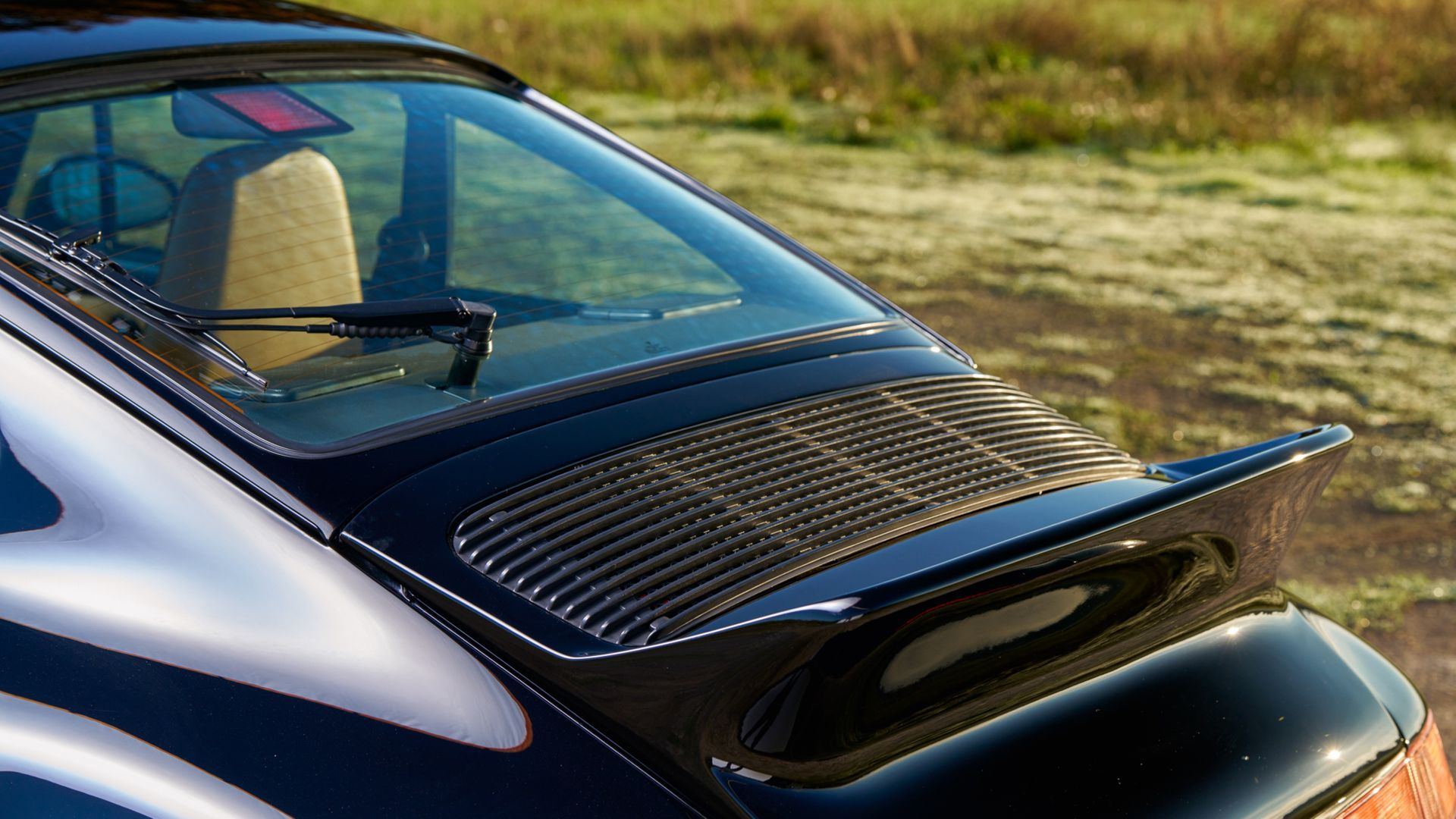 Black Porsche 911 rear, with spoiler, windshield, and air vent. Beige interior, grass background.